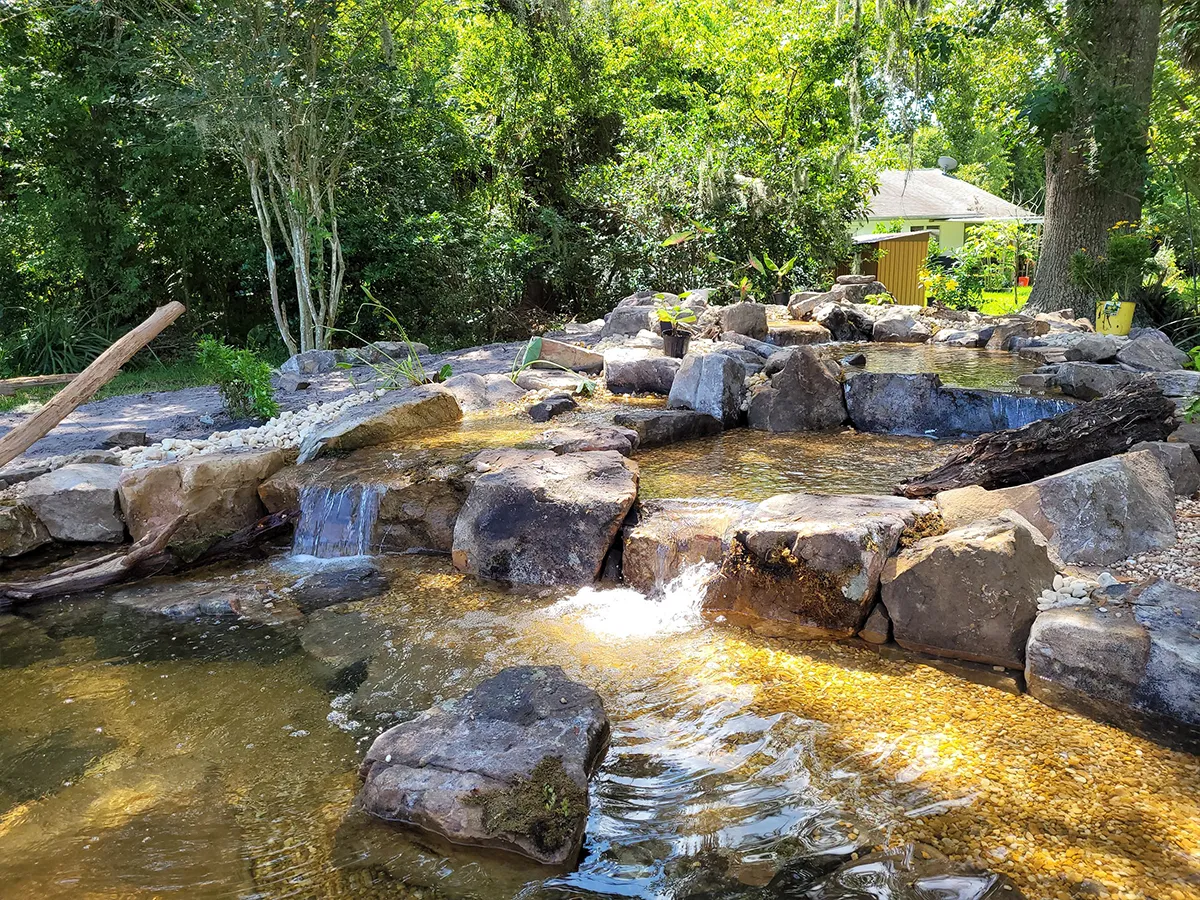 Natural garden pond with flowing water over rocks, surrounded by greenery and trees in a serene backyard setting.
