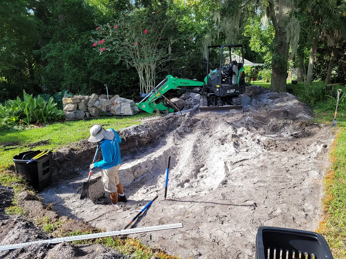 Excavation crew preparing a site for a backyard pond, using heavy equipment and manual tools.